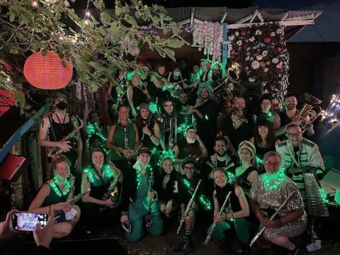 40 or so members of the Rude Mechanical Orchestra wearing green, black, and glitter hold their instruments and pose at Rubulad after their 18th anniversary show in June 2022.